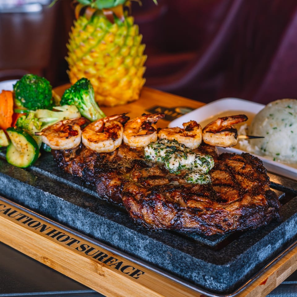 Grilled steak with herb butter, shrimp skewers, and mixed vegetables on a hot stone platter.