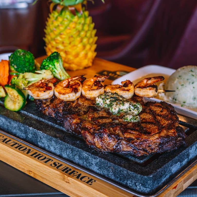 Grilled steak with herb butter, shrimp skewers, and mixed vegetables on a hot stone platter.