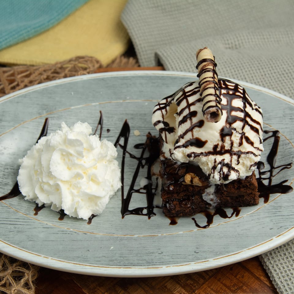 A brownie with vanilla ice cream, chocolate drizzle, a wafer roll, and whipped cream on a plate.