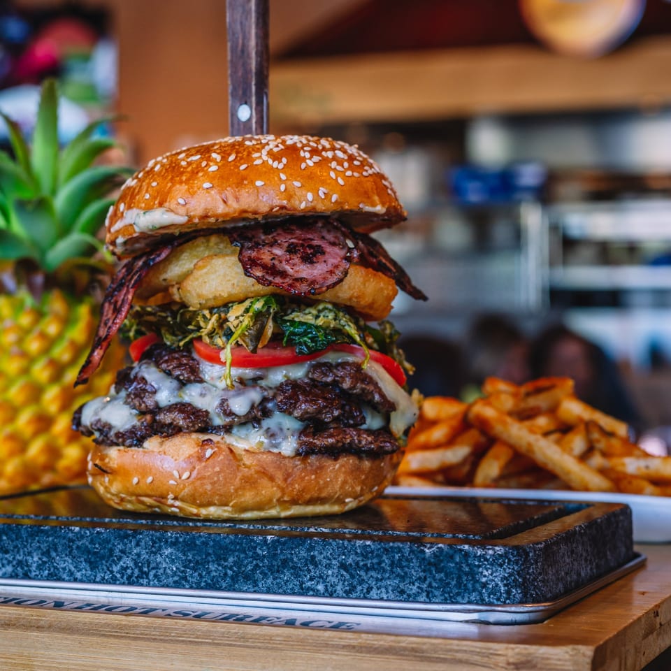 A large Peri Peri Original burger with multiple patties, bacon, and onion rings, served with fries.